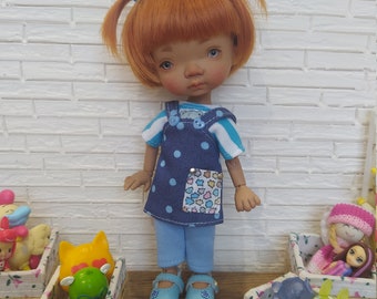 May include: A doll with ginger hair styled in two pigtails with blue flower clips, wearing a blue and white striped shirt under a navy blue polka dot overall dress with a multi-colored star pocket, light blue pants, and matching blue shoes.