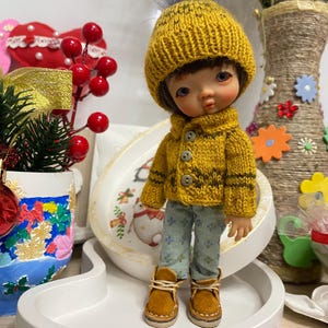 May include: A doll wearing a mustard yellow knit hat and matching sweater with brown accents. The doll is also wearing blue floral print pants and brown boots. The doll is standing on a white tray.