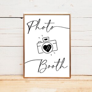 May include: A white sign with black text and a black and white illustration of a camera with a heart in the viewfinder. The sign reads "Photo Booth".