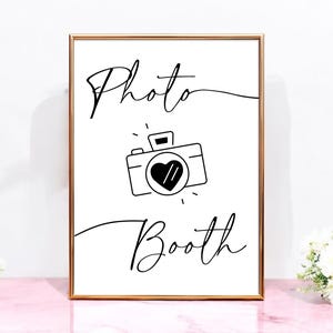 May include: A white sign with a gold frame that says "Photo Booth" in black cursive writing. A black and white illustration of a camera with a heart in the lens is above the text.