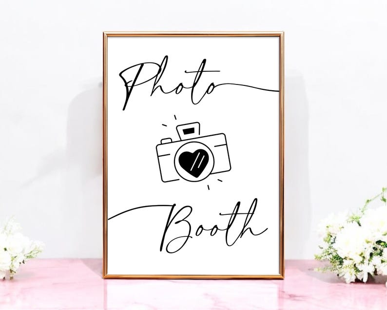 May include: A white sign with a gold frame that says "Photo Booth" in black cursive writing. A black and white illustration of a camera with a heart in the lens is above the text.