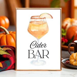 May include: A framed watercolor print with a gold border, featuring a cider cocktail with an apple slice garnish. The words "Cider BAR" are written in elegant black script. Pumpkins and a mug are in the background, creating a fall theme.
