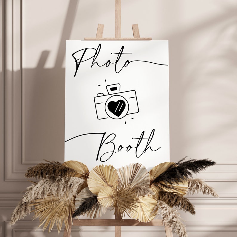May include: A white sign with black text and a camera icon that says "Photo Booth". The sign is on an easel and is surrounded by dried pampas grass.