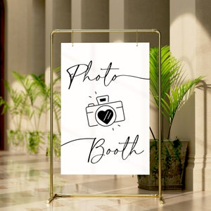 May include: A white sign with black text and a camera icon that says "Photo Booth". The sign is hanging from a gold metal stand.