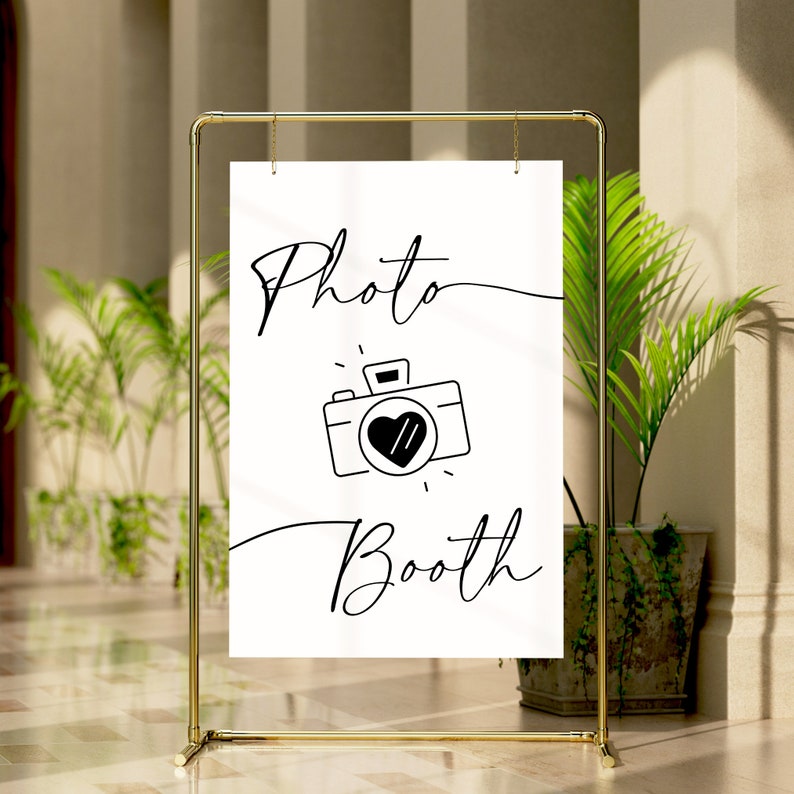 May include: A white sign with black text and a camera icon that says "Photo Booth". The sign is hanging from a gold metal stand.