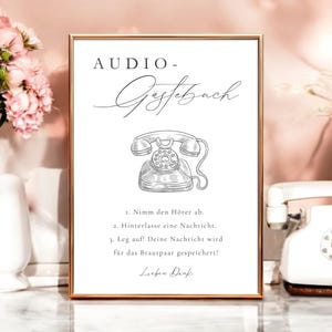 以下が含まれることがあります： 金色の縁取りのある額入りの看板には、「AUDIO - Gastebuch」という言葉とドイツ語での指示が書かれています。 ヴィンテージの電話のイラストがタイトルの下に配置されています。 看板はピンクの花と白い花瓶に囲まれています。