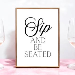 Puede incluir: Una impresión enmarcada con las palabras "Sip AND BE SEATED" en escritura negra y letras de bloque. El marco dorado se encuentra sobre una superficie rosa, con dos copas de vino a cada lado. Las copas están llenas de un líquido rosa claro.