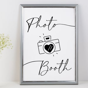 May include: A black and white printable sign with the text "Photo Booth" in a cursive font. A simple camera icon with a heart in the viewfinder is centered below the text.