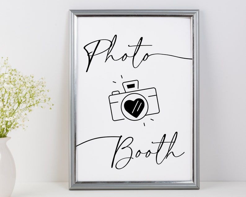 May include: A black and white printable sign with the text "Photo Booth" in a cursive font. A simple camera icon with a heart in the viewfinder is centered below the text.