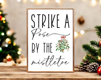 May include: A white printable sign with black text that reads "Strike a Pose By The Mistletoe". A watercolor illustration of a sprig of mistletoe with red berries hangs from the text.
