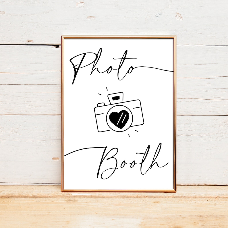 May include: A white sign with black text and a black and white illustration of a camera with a heart in the viewfinder. The sign reads "Photo Booth".