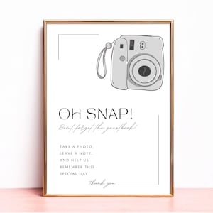 May include: A rectangular sign in a gold frame featuring a line drawing of an instant camera and the text: OH SNAP! Don't forget the guestbook! TAKE A PHOTO, LEAVE A NOTE, AND HELP US REMEMBER THIS SPECIAL DAY Thank you.