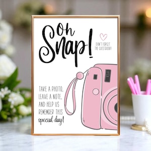 May include: A rectangular sign in a gold frame featuring the text Oh Snap! Don't forget the guestbook! and Take a photo, leave a note, and help us remember this special day! with a pink camera illustration on a white background.