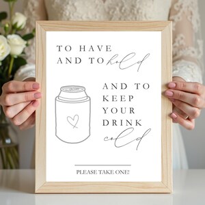 May include: A light wood frame containing a white sign with black text that reads: TO HAVE AND TO hold AND TO KEEP YOUR DRINK cold. Below the text is a line drawing of a beverage can cooler with a small heart outline and the text PLEASE TAKE ONE!
