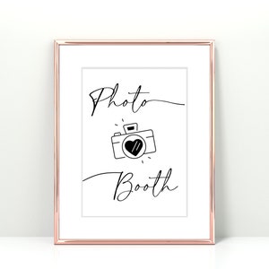 May include: A white sign with a black frame and the text "Photo Booth" in a cursive font. A black and white illustration of a camera with a heart in the viewfinder is below the text.