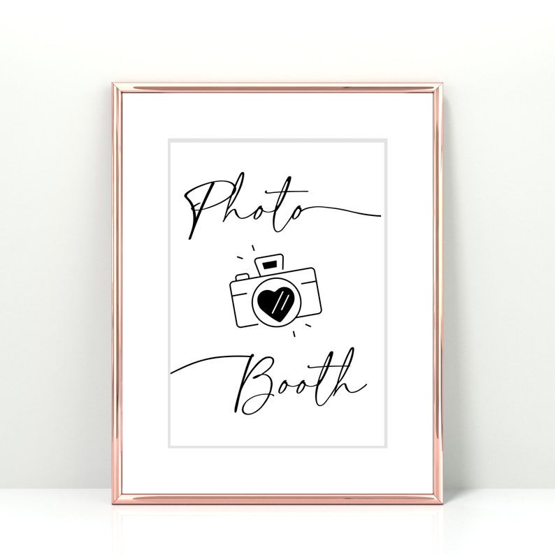 May include: A white sign with a black frame and the text "Photo Booth" in a cursive font. A black and white illustration of a camera with a heart in the viewfinder is below the text.