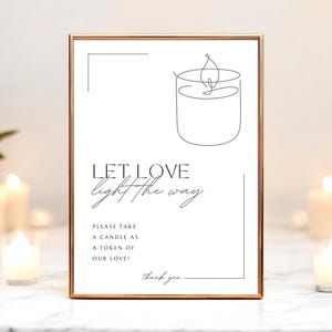 Puede incluir: Un letrero enmarcado con borde dorado presenta el texto "LET LOVE light the way." Debajo, se lee "PLEASE TAKE A CANDLE AS A TOKEN OF OUR LOVE!" Un dibujo lineal de una vela está en la parte superior. El letrero está rodeado de velas encendidas.