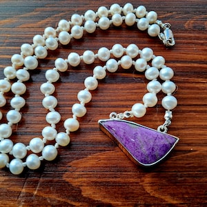 May include: A white pearl necklace with a purple gemstone pendant. The necklace is made of small, round pearls and has a silver clasp. The pendant is a triangular shape with a smooth, polished surface.