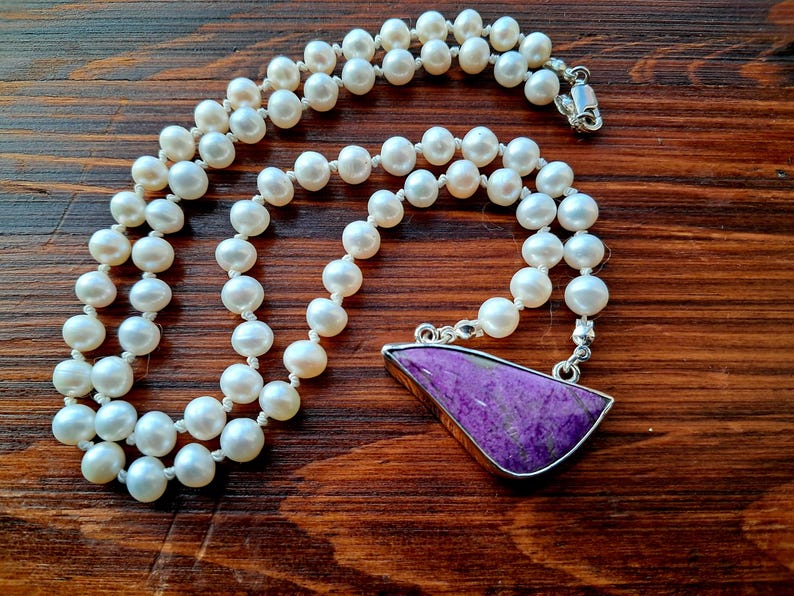 May include: A white pearl necklace with a purple gemstone pendant. The necklace is made of small, round pearls and has a silver clasp. The pendant is a triangular shape with a smooth, polished surface.