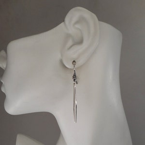 May include: Silver dangle earrings with a small silver charm at the top. The earrings are hanging from a mannequin's ear.