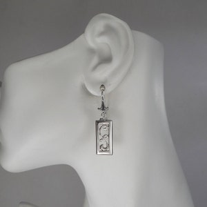 May include: Silver dangle earrings with a rectangular pendant featuring a delicate, intricate scroll design.
