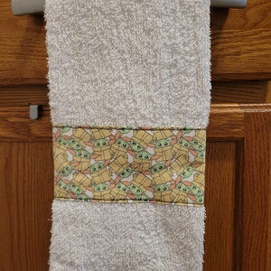 May include: White kitchen towel with a yellow and orange border featuring a repeating pattern of green cartoon characters with big eyes. The characters are wearing brown hats and are inside boxes.