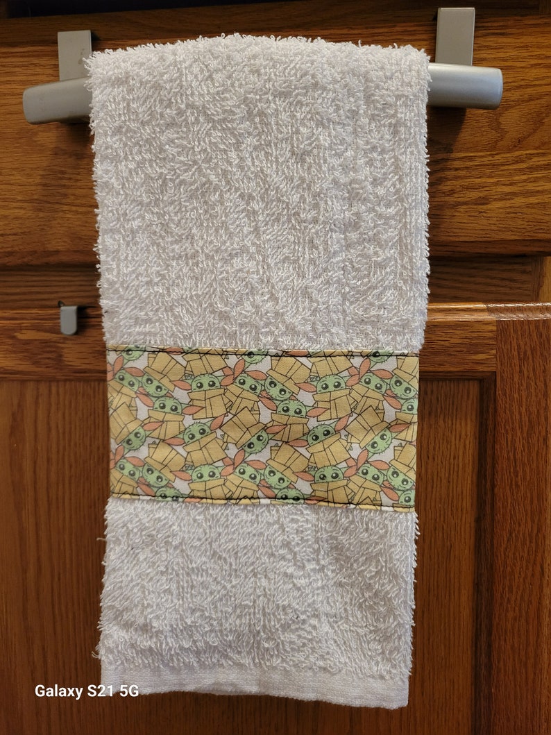 May include: White kitchen towel with a yellow and orange border featuring a repeating pattern of green cartoon characters with big eyes. The characters are wearing brown hats and are inside boxes.