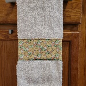 May include: White hand towel with a green and yellow pattern of Baby Yoda characters in boxes. The towel is hanging on a silver towel rack.