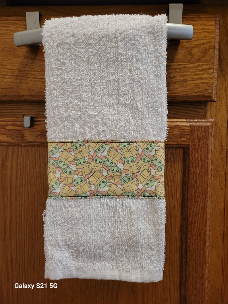 May include: White hand towel with a green and yellow pattern of Baby Yoda characters in boxes. The towel is hanging on a silver towel rack.