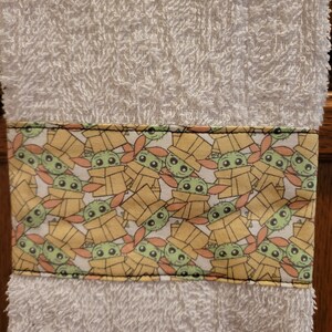 May include: White hand towel with a green and yellow border featuring a repeating pattern of a cartoon character with large eyes. The character is wearing a brown hooded robe and is surrounded by yellow boxes.