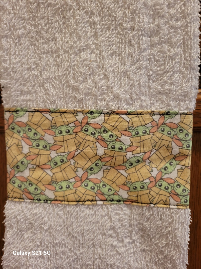 May include: White hand towel with a green and yellow border featuring a repeating pattern of a cartoon character with large eyes. The character is wearing a brown hooded robe and is surrounded by yellow boxes.