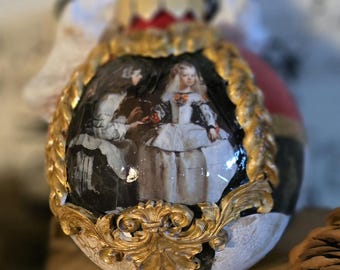 May include: An egg-shaped decorative ornament featuring a framed painting of two figures in historical attire. The frame is gold-colored with ornate detailing. The ornament has a black and white checkered pattern at the base.