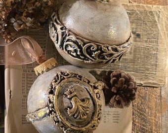 May include: Two antique-style Christmas ornaments with gold accents. One ornament features an ornate, swirling design, while the other has a gold emblem. Both are cream-colored with gold caps and ribbons, resting on aged paper.