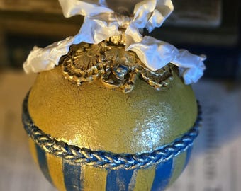 May include: A decorative, round ornament with a yellow and blue striped pattern. The ornament has a gold-colored top with a white ribbon and a textured, crackled finish. A blue rope-like trim separates the yellow and blue sections.