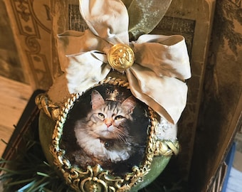 May include: A decorative Christmas ornament featuring a portrait of a cat wearing a crown. The ornament is green and gold, with a gold frame around the cat's portrait. A cream-colored bow and gold button adorn the top. The ornament is placed in a wooden box.