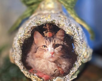 May include: A decorative bell-shaped ornament featuring a painted portrait of a cat. The ornament has a gold and cream-colored exterior with a green bow at the top. The cat portrait is framed within the bell, resting on a stack of old books.