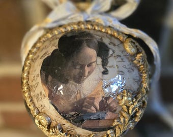 May include: An ornate, egg-shaped ornament featuring a vintage portrait of a woman writing. The ornament has a gold-colored filigree frame and a cream-colored background with handwritten script. A cream and gold ribbon is tied at the top.
