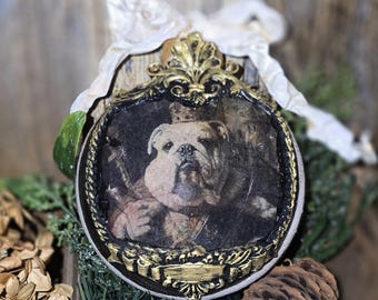 May include: An oval-shaped ornament featuring a regal bulldog portrait within a black and gold ornate frame. A white ribbon and greenery accent the ornament, which is set against a rustic wooden backdrop. A pinecone and dried flowers are also visible.