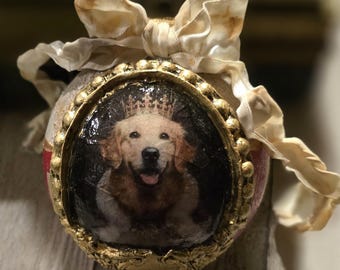 May include: An ornate, decorative ornament featuring a framed image of a golden retriever wearing a crown. The ornament has a gold-colored frame and is adorned with a cream-colored ribbon. The background is blurred, suggesting a focus on the ornament.