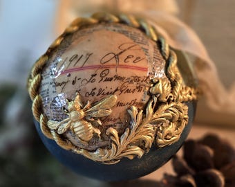 May include: An ornate, egg-shaped ornament with a dark blue base and a gold-colored, textured design. The top features a collage of vintage paper with handwritten text and a gold bee. A gold rope-like trim encircles the top.