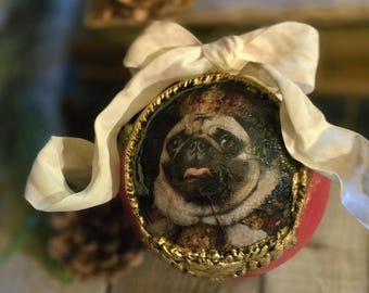 May include: A red and gold Christmas ornament featuring a pug wearing a crown. The ornament has a gold trim and is topped with a white ribbon bow. The background is blurred, with dried flowers visible.