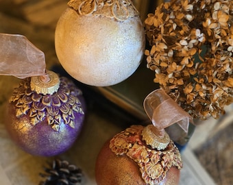 May include: Three decorative Christmas ornaments in shades of purple, gold, and bronze. Each ornament has a textured surface and is topped with a gold cap and a sheer ribbon. A dried flower arrangement and a pine cone are also visible.