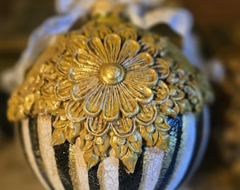 May include: A decorative ornament with a gold floral design on top and black and white stripes on the bottom. The ornament has a white ribbon and is likely for home decor.