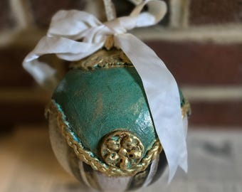 May include: A decorative Christmas ornament with a teal and gold design. The ornament has a gold trim and a white ribbon bow. The ornament is hanging in front of a brick wall.