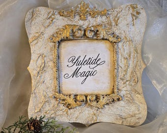May include: Ornate, square picture frame with a distressed, off-white finish and gold accents. The frame features the words "Yuletide Magic" in elegant script. A sprig of greenery and a pinecone are in the lower left corner.