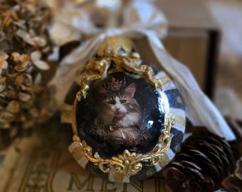 May include: Ornate Christmas ornament featuring a regal cat portrait within a gold-framed oval. The ornament has a black and gold striped pattern, topped with a white and gold ribbon. The text "CHARLES DICKENS" is visible on a book in the background.