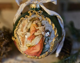 May include: A decorative Christmas ornament with a green base and a detailed image of a woman holding a child, framed with gold trim. The ornament has a white ribbon for hanging.