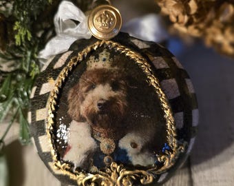May include: A round Christmas ornament featuring a portrait of a dog wearing a crown. The ornament has a black and white checkered pattern and is trimmed with gold. A gold button with a crown is at the top, with a white ribbon.