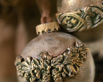 May include: Close-up of two decorative Christmas ornaments. The ornaments are round with a textured, metallic finish. One ornament features a gold-colored leaf and pinecone design around the bottom. The other ornament has a similar design.