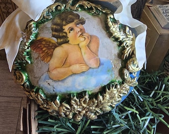May include: A vintage-style ornament featuring a cherubic angel on a cloud, framed by gold and green embellishments. A cream-colored ribbon and gold button add to the antique aesthetic. The ornament is placed on a wooden surface.